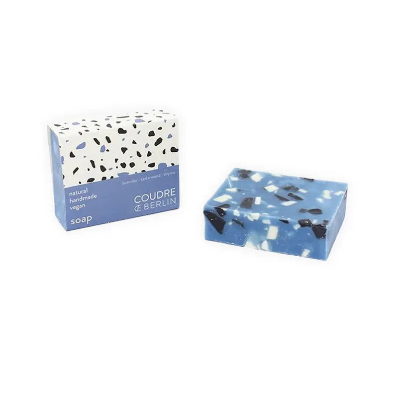 Ein blau-weißes handgefertigtes Seifenstück - Bluestone von Coudre Berlin ruht neben der passenden weißen Schachtel mit schwarzen Sprenkeln, die mit "COUDRE BERLIN natural handmade vegane Seife" beschriftet ist.