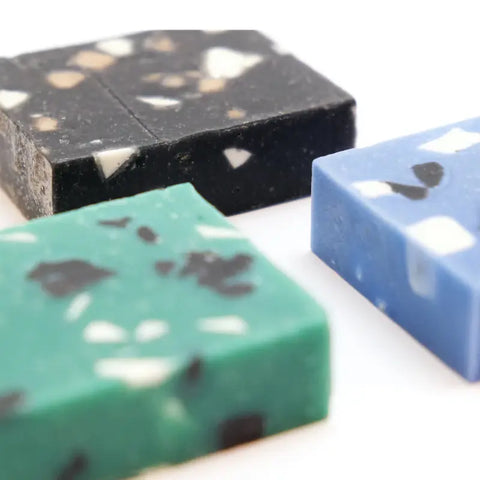 Drei rechteckige, handgefertigte Seifenstücke - Bluestone Bars von Coudre Berlin in Schwarz, Blau und Grün mit weißen und dunklen Sprenkeln sind auf einer weißen Fläche angeordnet und zeigen ihre handwerkliche Textur und ihre lebendigen Farben.