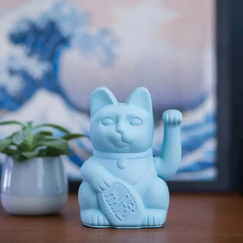 Eine Donkey Products Lucky Cat - Winkekatze hellblau Figur sitzt auf einer hölzernen Oberfläche neben einer kleinen Topfpflanze, mit einem sanft verschwommenen Wellenbild im Hintergrund.