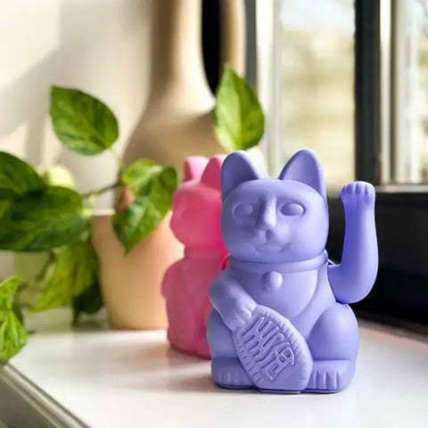 Die Lucky Cat - Winkekatze lila von Donkey Products, eine lila Maneki Neko mit erhobener Pfote, sitzt auf einer Fensterbank und versprüht Glücksbringer-Vibes, mit einer rosa Katzenfigur und einer Blattpflanze im Hintergrund.
