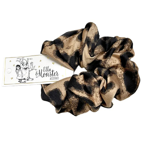 Ein hellbraunes und schwarzes gemustertes Leo Scrunchie von Ella & Monster mit einem weißen Anhänger mit einer Cartoon-Illustration von drei Figuren.