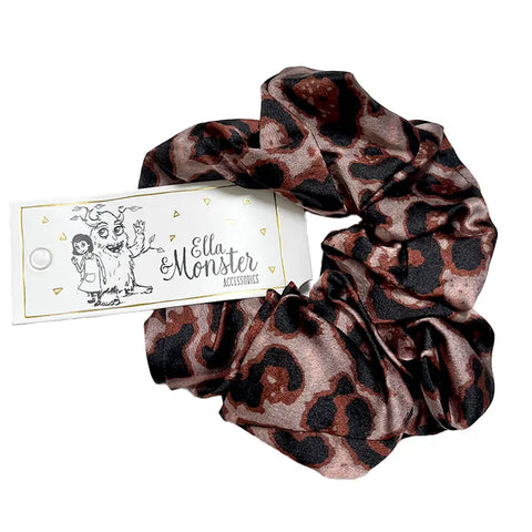 Das Leo Scrunchie von Ella & Monster ist ein seidiges, braun-schwarzes Haarschmuckstück mit Leopardenmuster und einem Logo-Anhänger, der ein Cartoon-Mädchen und ein Monster zeigt - das perfekte Haargummi für alle Fälle.