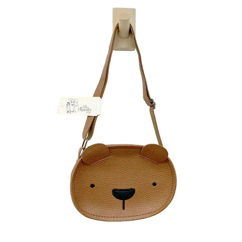 Die Tasche "Teddy" Kunstleder braun von Ella & Monster, eine niedliche braune Kindertasche in Bärenform mit runden Ohren und schwarzen Details, hängt an einem beigen Wandhaken. Sie verfügt über einen sicheren Reißverschluss und hat ein weißes Etikett am Gurt.