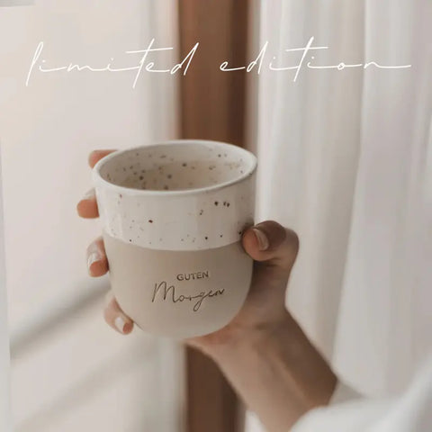 Eine Hand hält den Eulenschnitt Limitierter Becher "Guten Morgen" aus Steingut, beige-weiß gesprenkelt mit "limited edition" in Schreibschrift - ein perfektes Geschenk in sanftem Licht.
