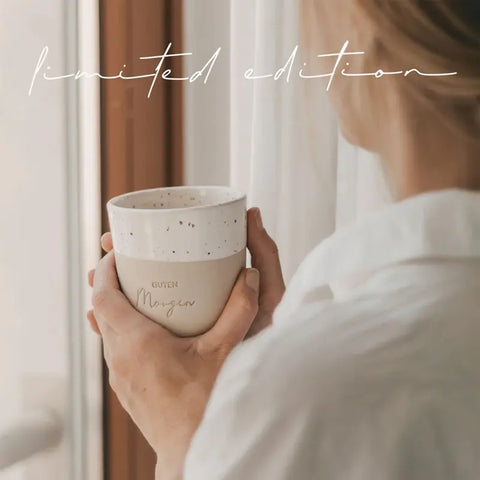 Eine Person in einem weißen Hemd hält den Eulenschnitt Limitierter Becher "Guten Morgen" aus Steingut in der Nähe eines Fensters mit transparenten Vorhängen. Darüber steht in Schreibschrift "Limited Edition", was ihn zu einer idealen Geschenkidee macht.