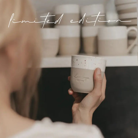 Eine blonde Person hält den Eulenschnitt Limitierter Becher "Lieblingsbecher" aus Steingut vor einem Regal mit ähnlichen Bechern, auf denen oben in Schreibschrift "Limited Edition" steht.