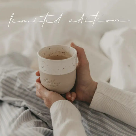 Eine Person in einem gemütlichen Pullover hält den mit Kaffee gefüllten Eulenschnitt Limitierter Becher "Lieblingsbecher" aus Steingut in der Hand, während sie auf einem Bett mit gestreifter und weißer Bettwäsche sitzt. Oben steht in Schreibschrift "Limitierte Auflage".