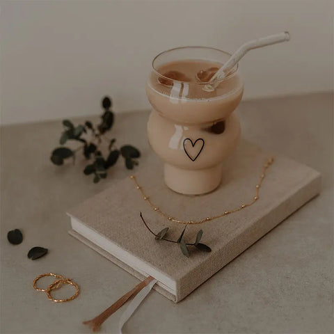 Das mit Eiskaffee gefüllte Double Bubble Glas "Herz" von Eulenschnitt ruht auf einem beigen Buch inmitten von Goldschmuck und Eukalyptusblättern - eine gemütliche, minimalistische Szene und das perfekte stilvolle Geschenk.