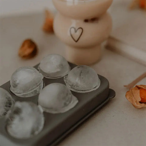 Eine Nahaufnahme einer Schale mit sechs runden Eiswürfeln auf einer hellen Oberfläche. Im Hintergrund sorgt das Eulenschnitt Double Bubble Glas "Herz" für Charme - perfekt für jedes stilvolle Ambiente oder als durchdachtes Geschenk.