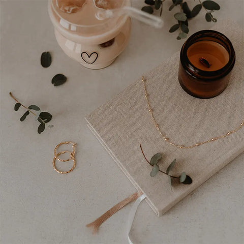 Ein beiges Stoff-Notizbuch mit einer zarten Goldkette und Eukalyptus ruht neben goldenen Ohrringen, einem braunen Kerzenglas und einem Eiskaffee aus dem Eulenschnitt Double Bubble Glas "Herz".