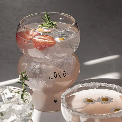 Ein rundes Eulenschnitt Double Bubble Glas "Love" enthält ein prickelndes Getränk mit Erdbeeren und Kräutern. In der Nähe stehen zwei weitere Gläser - eines in einem romantischen Glas mit Eis und Grünzeug, das andere mit schwebenden weißen Gänseblümchen.