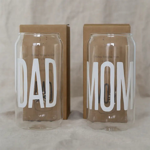 Zwei Gläser Hohes Trinkglas "Dad" von Eulenschnitt in Form von Getränkedosen mit den Aufschriften DAD und MOM stehen vor braunen Schachteln - ein perfektes Geschenk für Papa oder ein einzigartiges Geschenk für den Papa.