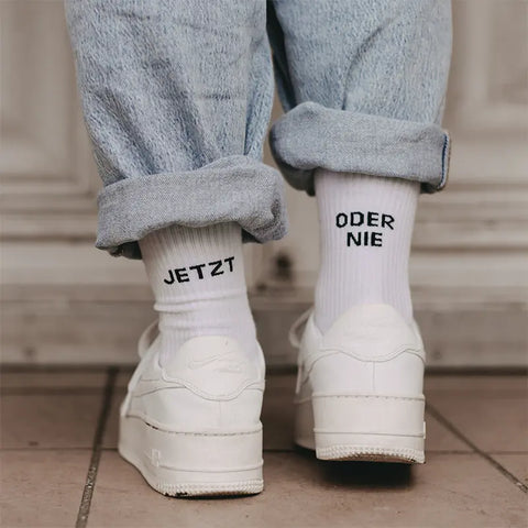 Eine Person trägt hochgekrempelte hellblaue Jeans, weiße Turnschuhe und Eulenschnitt Socken "Jetzt oder Nie" weiß mit schwarzer Schrift "JETZT" auf der einen und "ODER NIE" auf der anderen Socke.