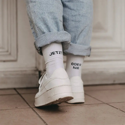 Eine Person in hellblauen Jeans mit Bündchen, weißen Turnschuhen und Eulenschnitt Socken "Jetzt oder Nie" weiß - mit "JETZT" auf der einen und "ODER NIE" auf der anderen Seite - steht auf einem gefliesten Boden.