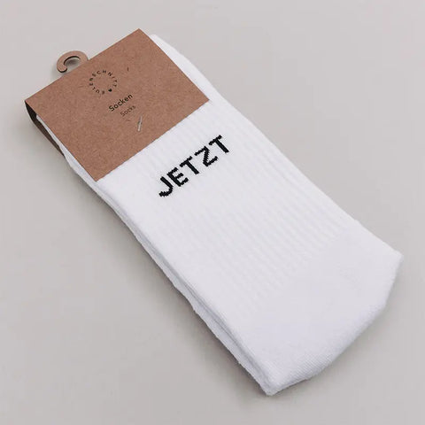 Ein Paar Eulenschnitt Socken "Jetzt oder Nie" in weiß, gefaltet mit einem braunen Kartonetikett; auf den Socken steht JETZT in fetten schwarzen Großbuchstaben.