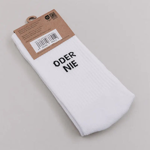 Ein Paar Socken "Jetzt oder Nie" in Weiß von Eulenschnitt, gefaltet unter einem braunen Kartonetikett mit Barcode, präsentiert auf einer hellgrauen Fläche.