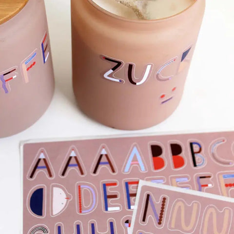 Zwei beigefarbene Gläser mit farbenfrohen Buchstaben-Aufklebern werden mit dem Stickerbogen - ABC (2 Bögen) von Halfbird mit Großbuchstaben-Aufklebern in verschiedenen Designs auf rosa Hintergrund präsentiert. Die Gläser sind aus umweltfreundlichem Recyclingpapier hergestellt.