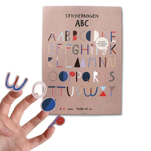 Eine Hand hält mehrere dekorative Alphabetsticker vor einem beigen Bogen mit der Aufschrift Stickerbogen - ABC (2 Bögen) von Halfbird, auf dem bunte Motive in Großbuchstaben auf Recyclingpapier gedruckt sind.
