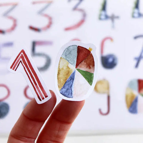 Zwei Finger halten Papierausschnitte: einer ist eine rot gestreifte Zahl aus dem Halfbird Stickerbogen - Zahlen (2 Bögen), der andere ist ein buntes, kreisförmiges Tortendiagramm. Im Hintergrund erscheinen verschwommene Zahlen in Aquarellfarben.