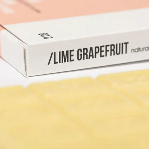 Eine Nahaufnahme einer weißen Hopfenschachtel mit der Aufschrift "Lime Grapefruit - Badeschokolade" in fetten schwarzen Buchstaben, die palmölfreie Naturkosmetik zeigt. Das 80g Gewicht und ein hellrosa Abschnitt erscheinen vor einem verschwommenen gelben Hintergrund.