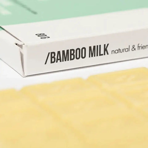 Eine Nahaufnahme einer Hopery Bamboo Milk - Badeschokolade Schachtel mit der Aufschrift natural & friendly, mit einem Teil einer beigen Schokoladentafel aus Kakaobutter - ideal für Fans von veganer Bade Schokolade - im Vordergrund sichtbar.
