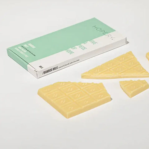 Eine Hopery Bamboo Milk - Badeschokolade Tafel in hellgrüner und weißer Verpackung sitzt hinter einer gebrochenen weißen Schokoladentafel auf einer weißen Fläche. Diese Badeschokolade wird mit cremiger Kakaobutter für ein luxuriöses Badeerlebnis hergestellt.