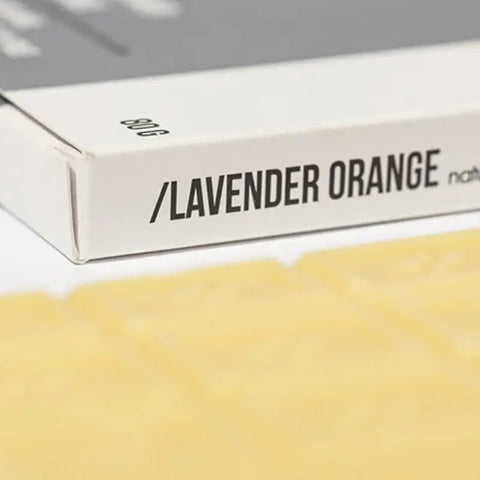 Eine Nahaufnahme einer weißen Schachtel mit der Aufschrift "Lavendel Orange - Badeschokolade" von Hopery zeigt einen Teil der gelblichen, rechteckigen Folie. Die 80g-Packung suggeriert ein wohlriechendes, beruhigendes Badevergnügen mit reichhaltigen Aromen.