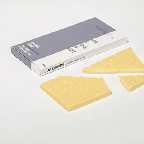 Eine teilweise zerbrochene Hopery Lavender Orange - Badeschokolade vegane weiße Schokolade mit Kakaobutter (ohne Palmöl) wird vor ihrer grau-weißen Verpackung auf einer weißen Fläche präsentiert.