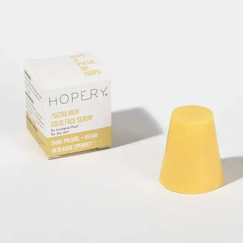 Ein solider gelber Kegel in Form eines Ultra Rich Face Serums für trockene Haut von Hopery steht neben der weiß-gelben Schachtel, die die Vorteile der veganen Hautpflege und die Unterstützung für trockene Haut auf der Verpackung hervorhebt.