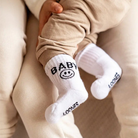 Ein Baby trägt Loones Socken "Baby" aus weicher Bio-Baumwolle, mit "BABY" und Smiley hinten sowie "LOONES" auf der Sohle. Es sitzt auf dem Schoß eines Erwachsenen in Beige.