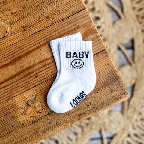 Ein Paar Loones Socken "Baby" aus Bio-Baumwolle liegt auf einem Holztisch. Die nachhaltigen weißen Socken zeigen "BABY", ein lächelndes Gesicht und das Wort "LOVER" bei den Zehen.