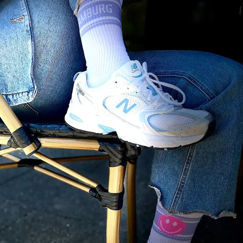 Eine Person in hellblauen New Balance Turnschuhen, blauen Jeans mit Bündchen und Loones Socken "HAPPY HAMBURG | Smiley" sitzt auf einem Klappstuhl und hat ein Bein über das andere geschlagen.