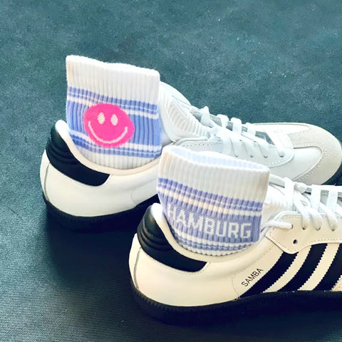 Ein Paar weiße Adidas-Samba-Turnschuhe mit schwarzen Streifen, die jeweils eine Loones-Socke "HAPPY HAMBURG | Smiley" tragen: Auf der einen ist ein rosa Smiley abgebildet, auf der anderen steht "HAMBURG" in weißer Schrift auf weicher Bio-Baumwolle.