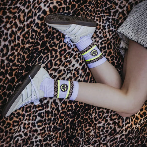 Eine Person liegt auf einer Decke mit Leopardenmuster und zeigt nur Beine und einen Teil eines Arms in einem Strickpullover. Sie trägt weiße Turnschuhe und Loones Socken "LEO Smiley | white" in hellem Lila mit gelben und schwarzen Streifen sowie einem Löwenemblem.
