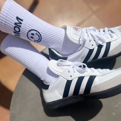 Eine Person in weißen Adidas Samba-Turnschuhen mit schwarzen Streifen und Loones Socken "Mom" - weiße, nachhaltige Damensocken mit MOM-Print und Smiley - steht auf einem gefliesten Boden und setzt einen verspielten, umweltfreundlichen Akzent zum klassischen Look.