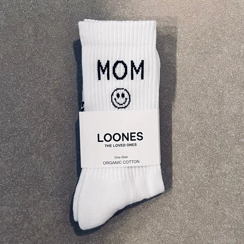 Ein Paar weiße Loones Socken "Mom" mit Smiley aus Bio-Baumwolle, nachhaltig und One Size, mit THE LOVED ONES-Label, liegt auf grauem Untergrund.