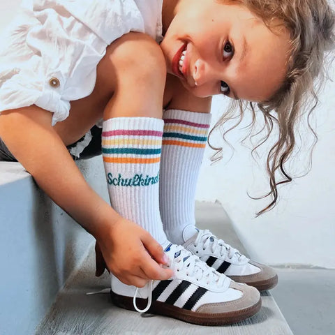 Lächelndes Kind im weißen Hemd aus Bio-Baumwolle und Loones Socken "Schulkind"-Petrol, das auf den Stufen sitzt und weiße Adidas-Turnschuhe mit schwarzen Streifen bindet - bereit für den Schulanfang.