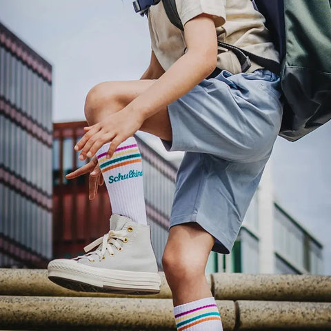 Ein Kind in hellblauen Shorts, weißen Turnschuhen und Loones Socken "Schulkind"-Petrol tritt mit einem Rucksack auf einen Steinvorsprung im Freien - bereit für den Schulanfang, im Hintergrund sind Gebäude zu sehen.