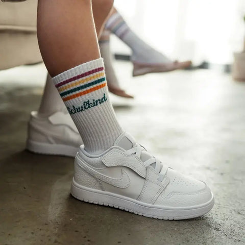 Jemand, der Loones Socken "Schulkind" in Petrol und weiße Turnschuhe trägt, sitzt in einem Haus. Bunte Streifen und die "Schulkind"-Stickerei sorgen für eine verspielte Note - perfekt für den Schulanfang. Eine weitere Person in ähnlichen Socken ist im Hintergrund zu sehen.