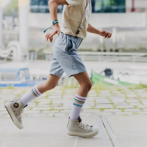 Ein Kind in Schuluniform und High-Top-Turnschuhen, das Loones Socken "Schulkind" petrol trägt, läuft im Freien auf einem gepflasterten Weg. Nur die untere Hälfte des Körpers des Kindes ist zu sehen, der Hintergrund ist leicht verschwommen.