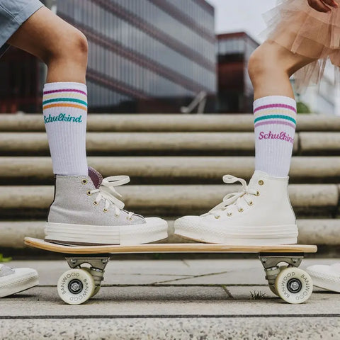Zwei Kinder stehen sich auf einem Skateboard gegenüber, beide tragen High-Top-Turnschuhe und Loones Socken "Schulkind"-Petrol und stehen auf einer Steintreppe im Freien mit modernen Gebäuden im Hintergrund.