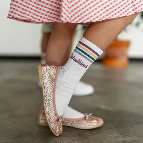 Nahaufnahme eines Kindes am Schulanfang, das glitzernde rosa Schuhe und Loones Socken "Schulkind"-Plaume mit Regenbogenstreifen und "Socksistkind"-Schriftzug trägt, gepaart mit einem rosa Rock mit Karomuster.