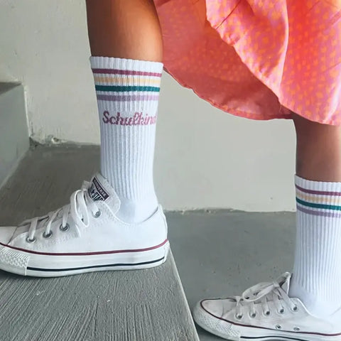 Ein Kind steht auf einer grauen Treppe in einem rosa gepunkteten Rock, trägt Loones Socken "Schulkind" Pflaume und weiße Turnschuhe - perfekt für den Schulanfang.