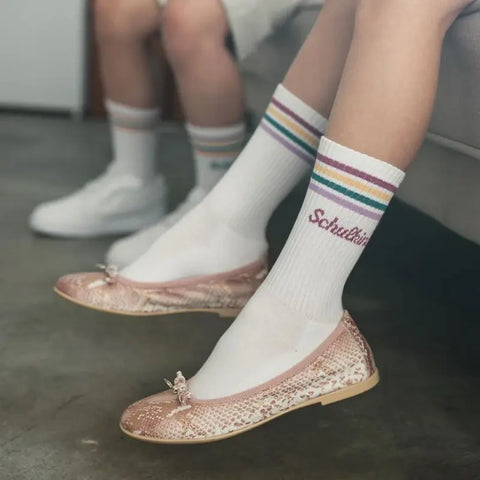 Zwei Menschen sitzen nebeneinander auf einer hellen Couch. Die eine trägt glitzernde rosa Ballerinas und Loones Socken "Schulkind" pflaume mit bunten Streifen, hergestellt aus Bio-Baumwolle. Die andere hat weiße Turnschuhe und Socken, bereit für den Schulanfang.
