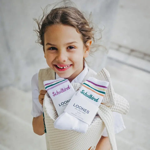 Ein lächelndes Kind mit lockigem Haar hält eine Loones Tragetasche mit zwei Paar Socken "Schulkind" pflaume. Die bunt gestreiften Bio-Baumwolle-Socken sind perfekt für den Schulanfang! Das Kind im weißen Hemd steht in einem Raum.