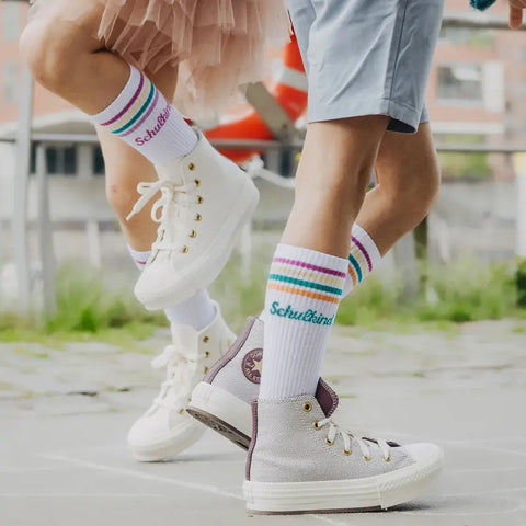 Zwei Kinder in weißen High-Tops und Loones Socken "Schulkind" Pflaume laufen im Freien - eines im rosa Tutu, das andere in hellblauen Shorts. Ideal für Schulanfang-Abenteuer mit bequemen Bio-Baumwolle-Socken!.