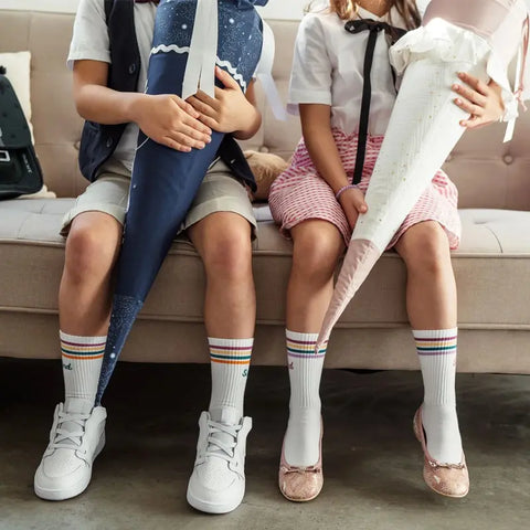 Zwei Kinder sitzen bei Schulanfang auf einer Couch, jedes mit einer Schultüte. Beide tragen Loones Socken "Schulkind" Pflaume - weiße Kniestrümpfe mit Regenbogenstreifen - kombiniert mit Shorts und Turnschuhen oder einem Rock und Flats.