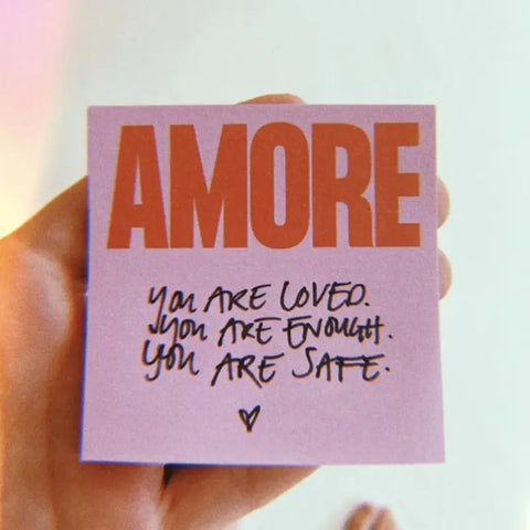Eine Hand hält die Navucko Haftnotizen "Amore" - Rosa/Rot, 50 Blatt, mit einem lila Zettel mit AMORE in fetter roter Schrift und handgeschriebenen Botschaften: Du wirst geliebt. Du bist genug. Du bist sicher, gekrönt von einem kleinen Herz.