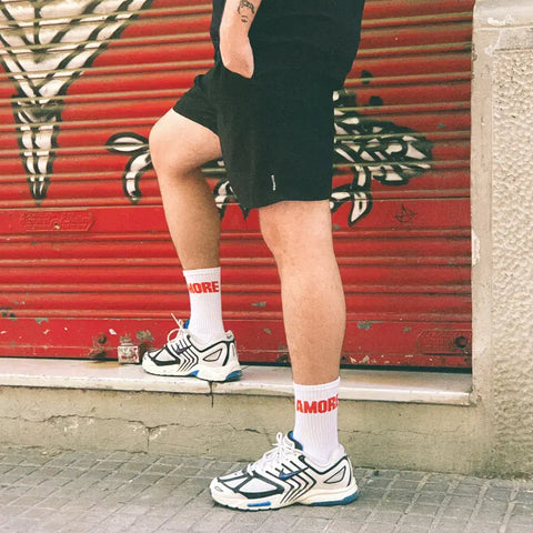 Eine Person in weißen Turnschuhen, Navucko-Socken "Amore" und schwarzen Shorts steht vor einem rot-silbernen Metalltor mit Graffiti. Nur ihre Beine sind sichtbar.