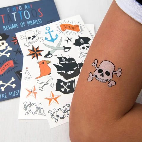 Ein Kinderarm mit einem schwarz-weißen Totenkopf aus Rex Londons Abwaschbare Tattoos - Piraten (2 Bögen). Im Hintergrund: Bögen mit Piratentattoos - Anker, Schiffe, Hüte, Sterne - perfekt für kleine Abenteurer!.